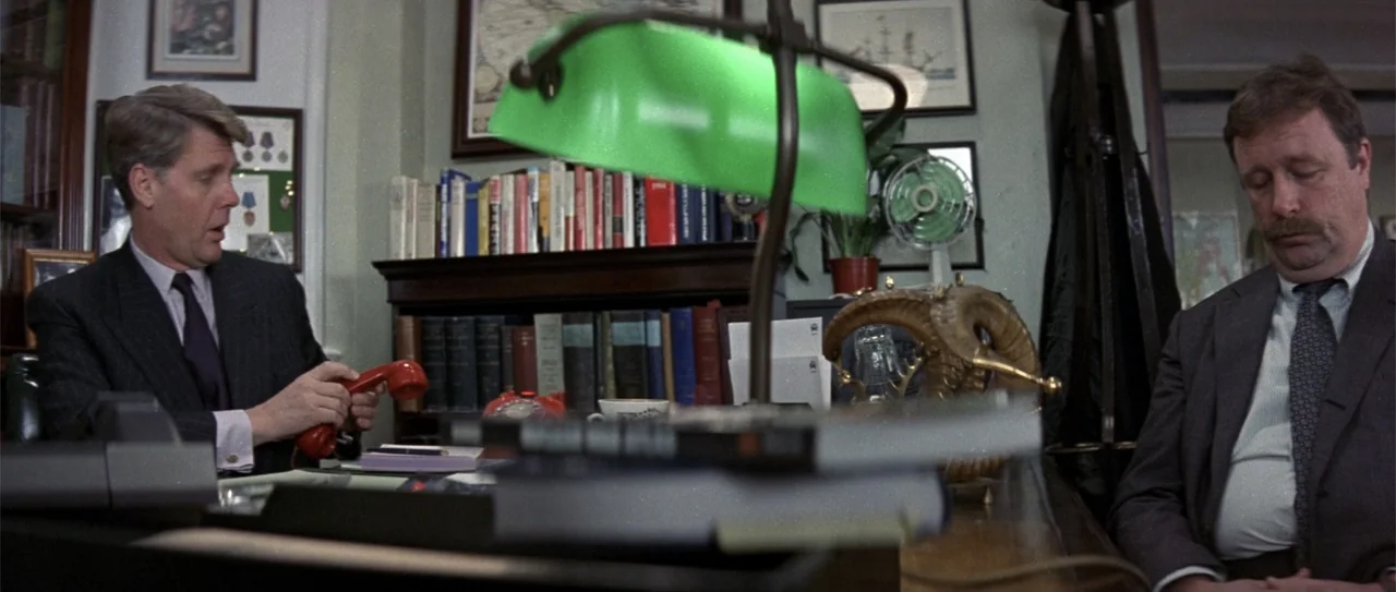 The Russia House (1990) movie still: medium — An wood-paneled office contains bookshelves, a desk with a green lamp, and framed pictures. Two men in…