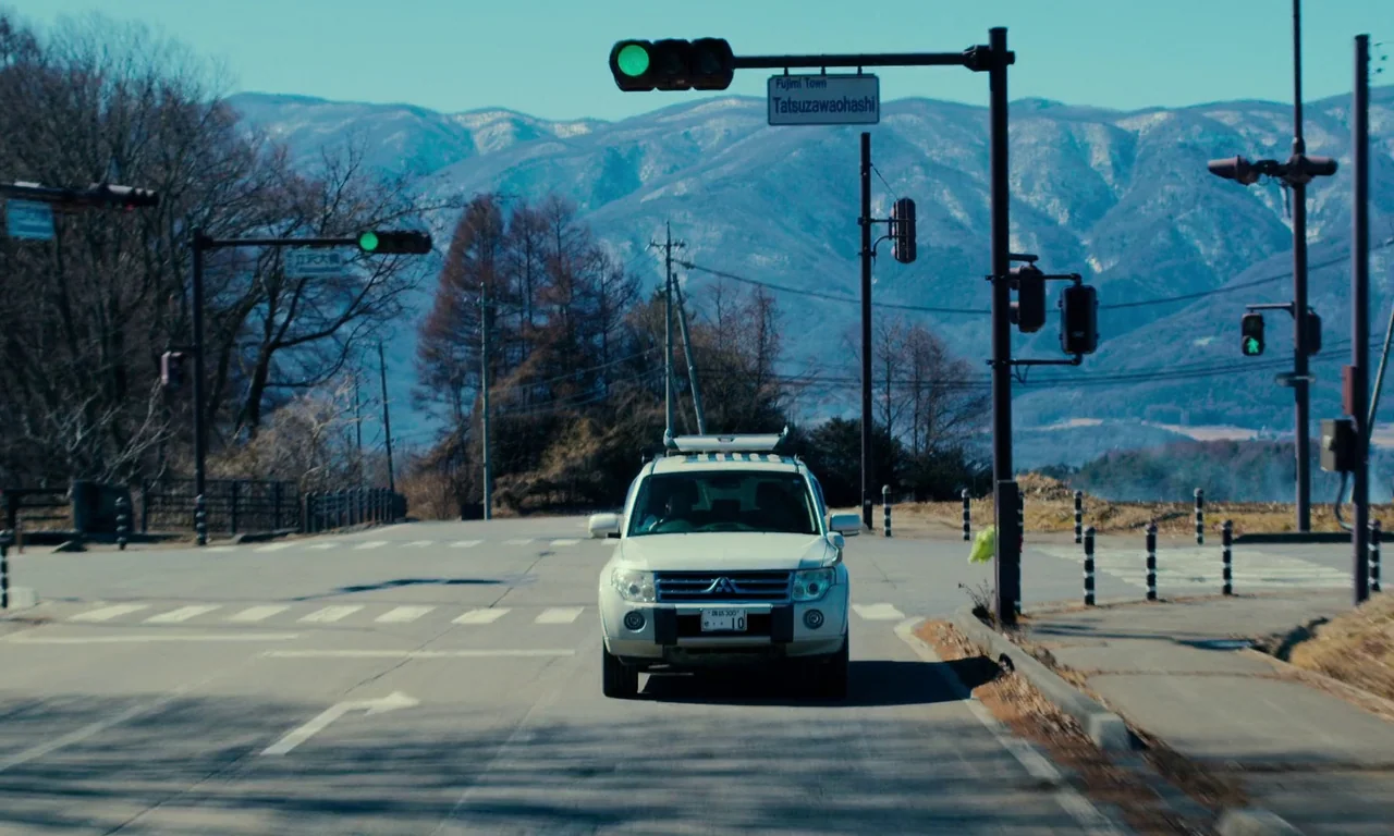 Evil Does Not Exist (2023) movie still: wide — A rural paved intersection features a white SUV driving toward the camera. In the background, rolling…
