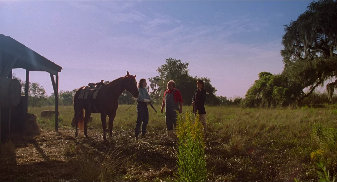 Hard Target (1993) movie still: wide — A grassy field with a wooden shed and mature trees under a clear sky. Three people stand in a line while a…