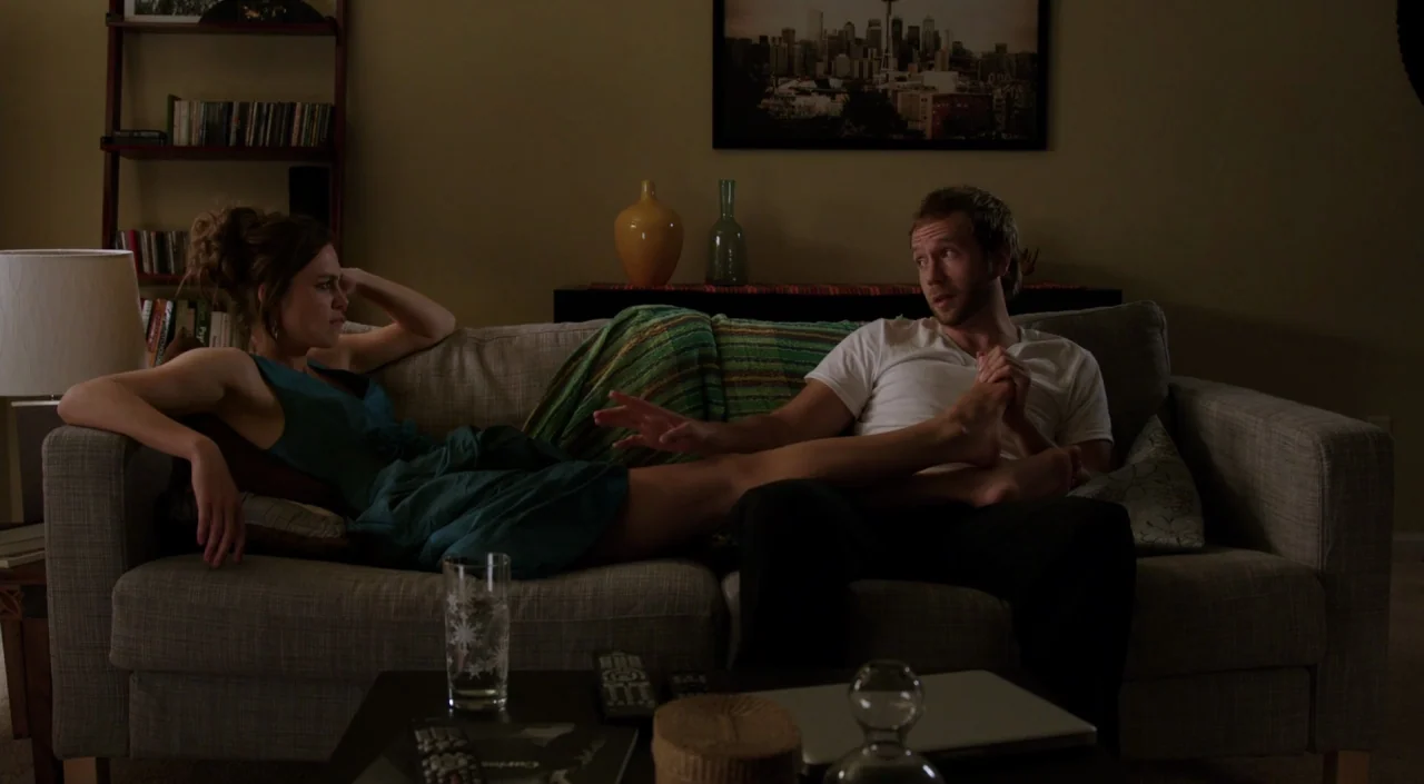 Laggies (2014) movie still: medium — A residential room features a couch against a wall with framed art and decor. The man and woman sit together on…