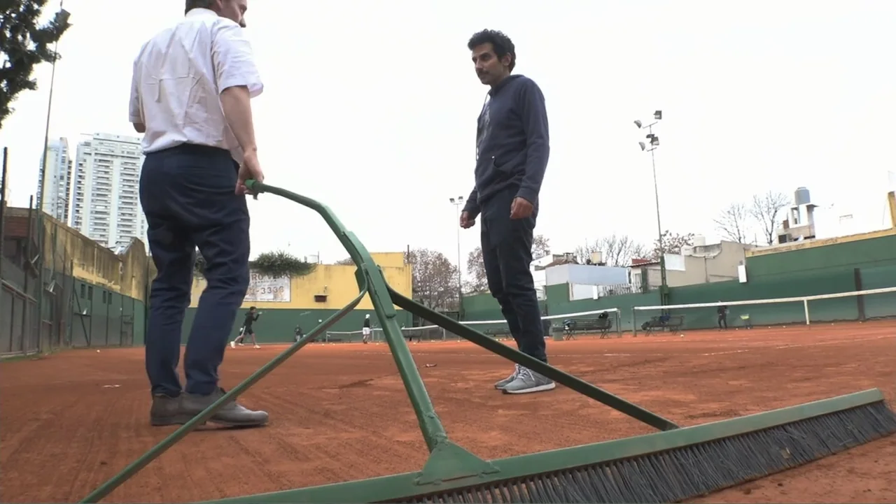 Smog in Your Heart (2022) movie still: fullbody — A clay tennis court features two men standing on the red dirt surface. One man holds a long metal…