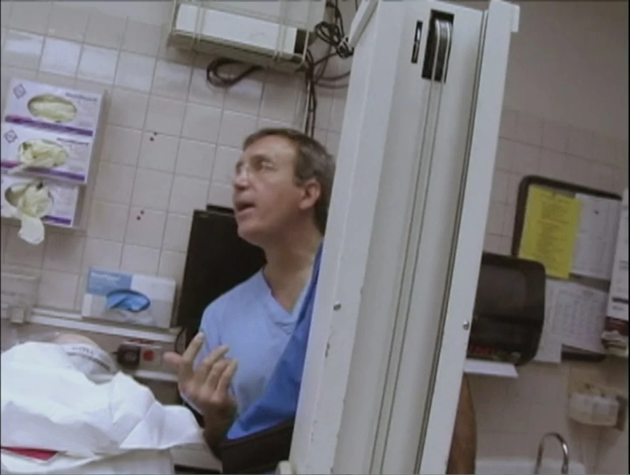 Addiction (2024) movie still: medium — A sterile clinical room features white tiled walls with medical supply boxes attached. A man wearing blue…