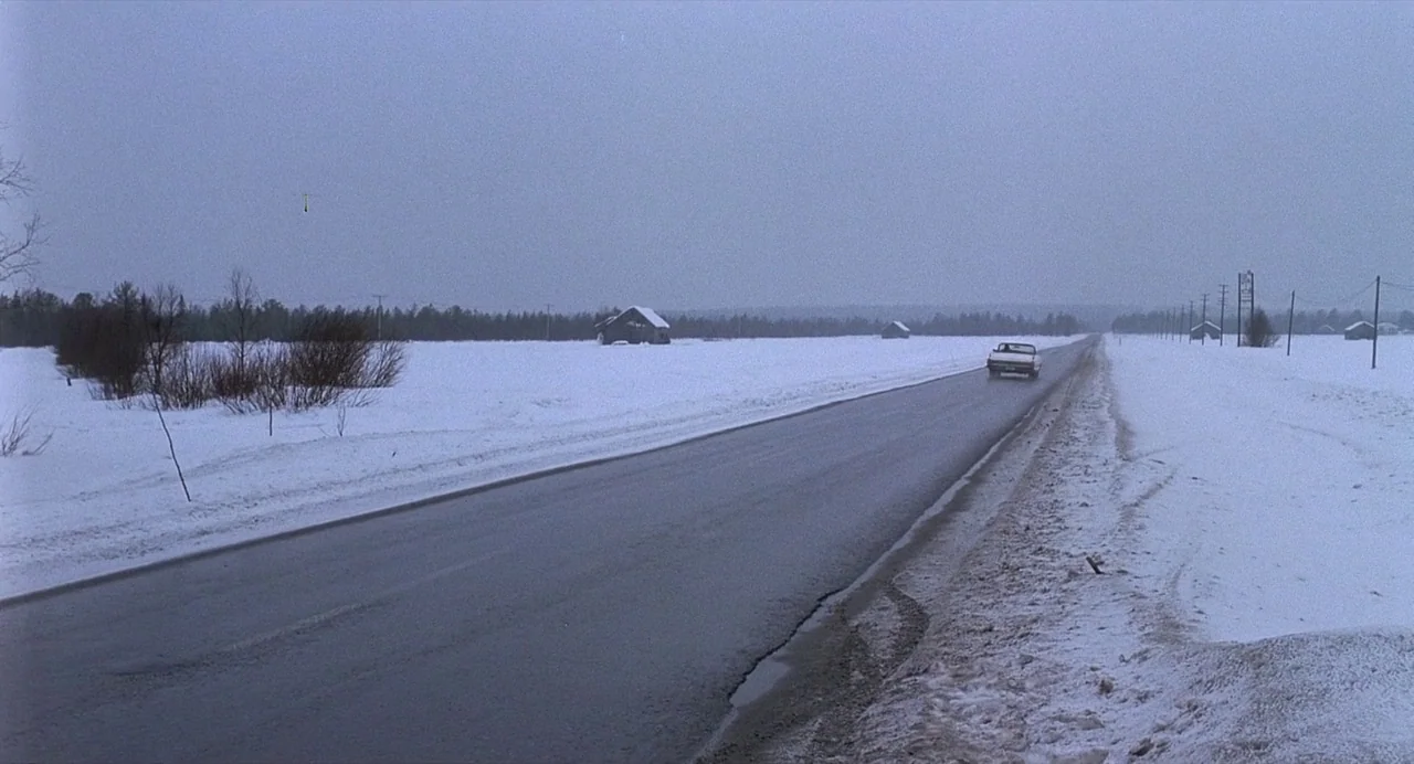 Ariel (1988) movie still: wide — A long, straight stretch of asphalt cuts through a flat, snow-covered rural landscape. A single car moves away from…
