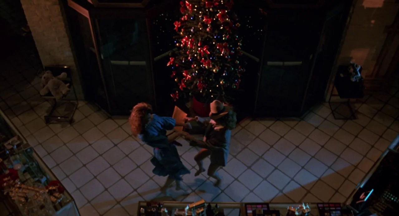 Night of the Comet movie still: wide — A department store interior features a large decorated Christmas tree on a tiled floor. A man and a woman in…