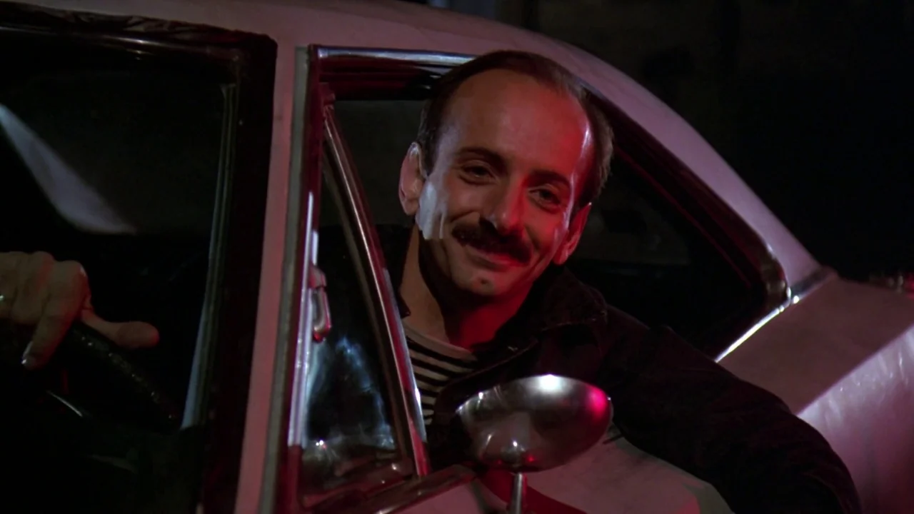 Friday the 13th: A New Beginning (1985) movie still: medium — A man sits inside a dark car at night under red and dim lighting. He smiles while…