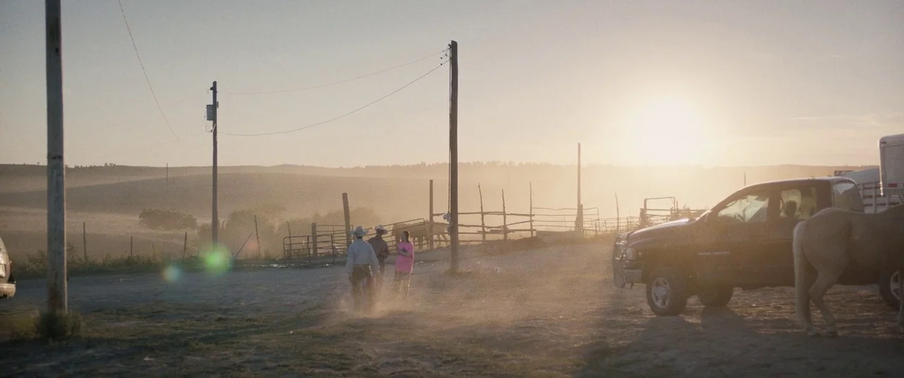 The Rider (2017) movie still: wide — A dusty dirt lot at a ranch during sunset with rolling hills in distance. Three people stand together near a…