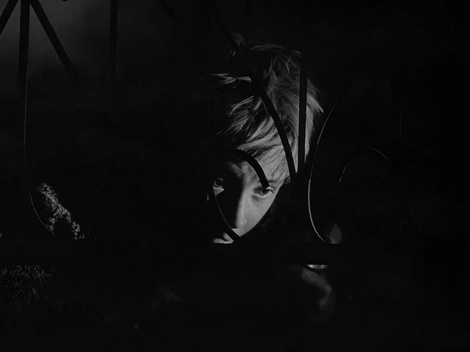 Ivan’s Childhood (1962) movie still: closeup — A dark outdoor space features an ornate black metal fence in the foreground. A young boy looks…