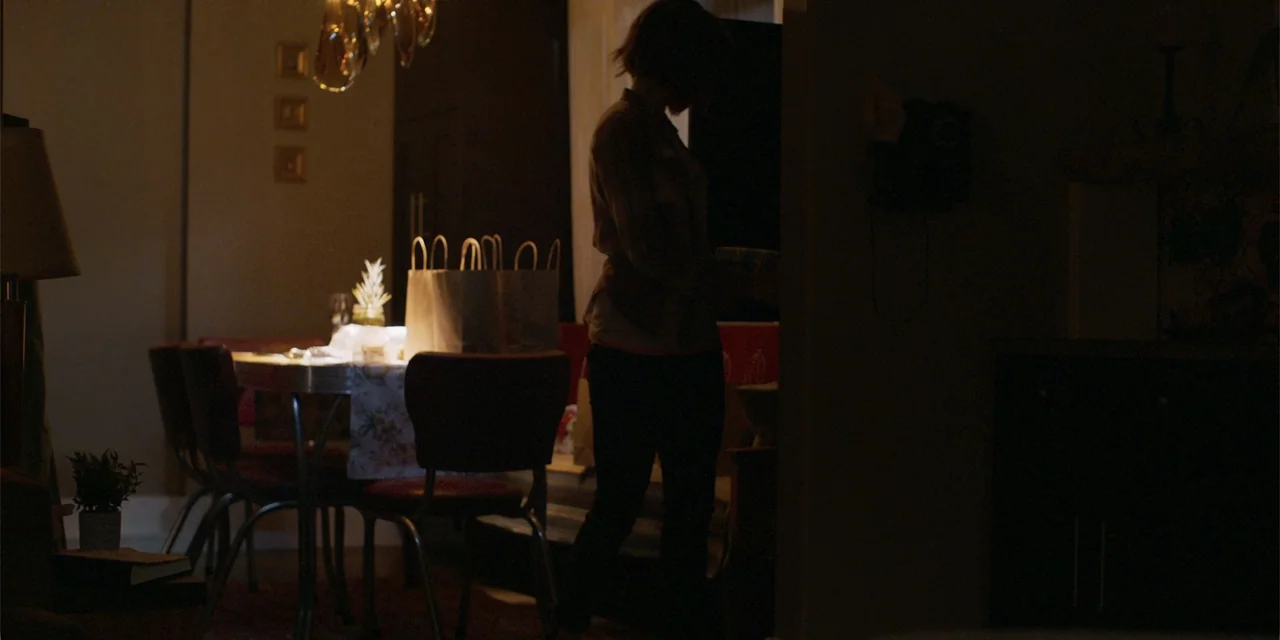 Demonic (2015) movie still: medium — A dim dining room contains a small table set with paper bags and decorative items. A woman stands near the…