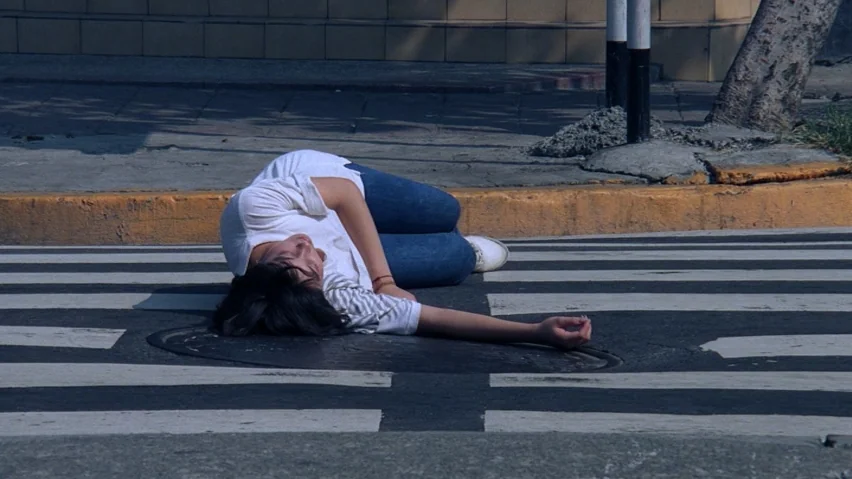 The Terrorizers (1986) movie still: medium — A crosswalk on an asphalt road with a circular metal manhole cover. A young woman lies motionless in a…