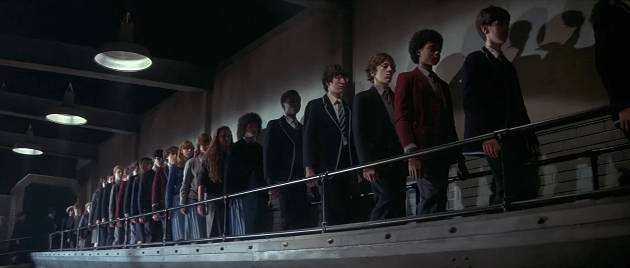 Pink Floyd The Wall (1982) movie still: wide — A long queue of students in uniform marches across a raised metal platform. The industrial space…