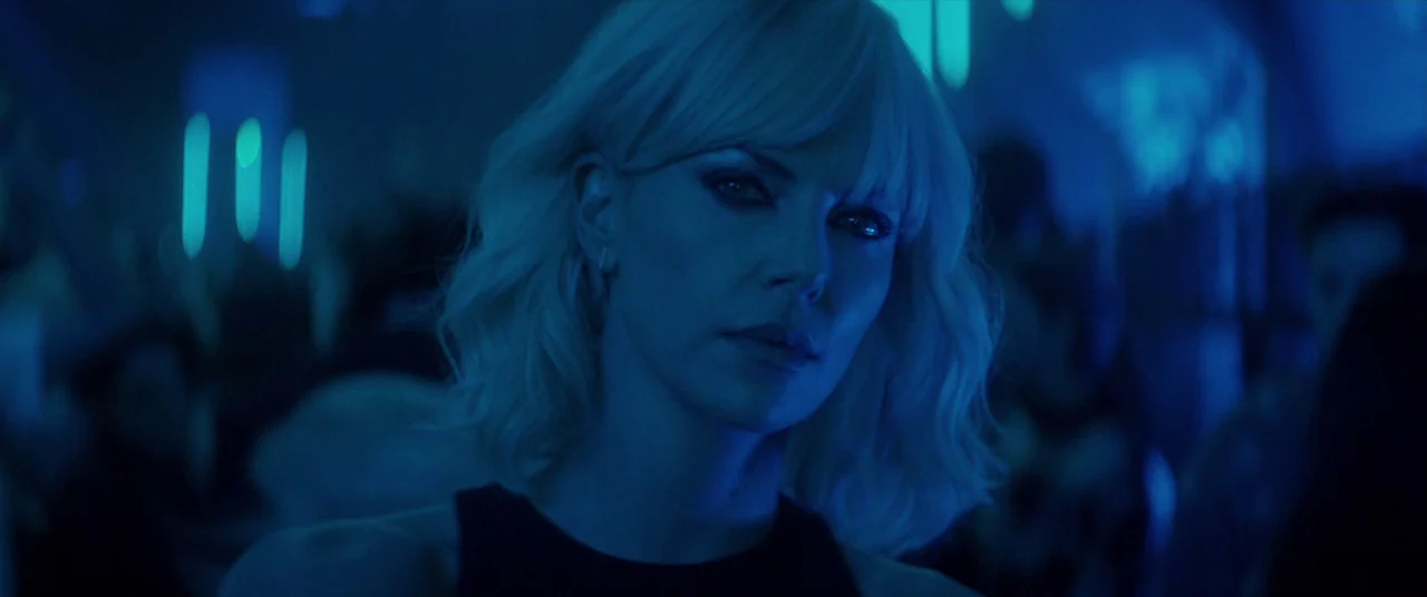 Atomic Blonde (2017) movie still: closeup — A dark, crowded interior space features vertical cyan light bars glowing against deep shadows. A blonde…