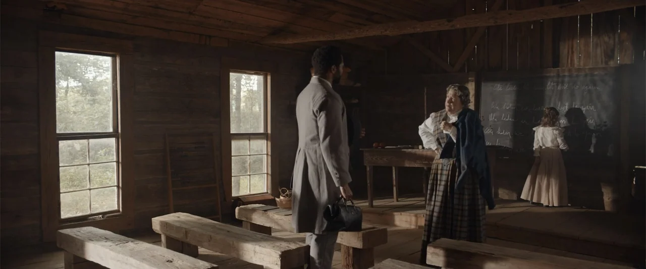 Ghosts of the Ozarks (2022) movie still: medium — A rustic one-room wooden building features exposed log walls, simple benches, and a large…