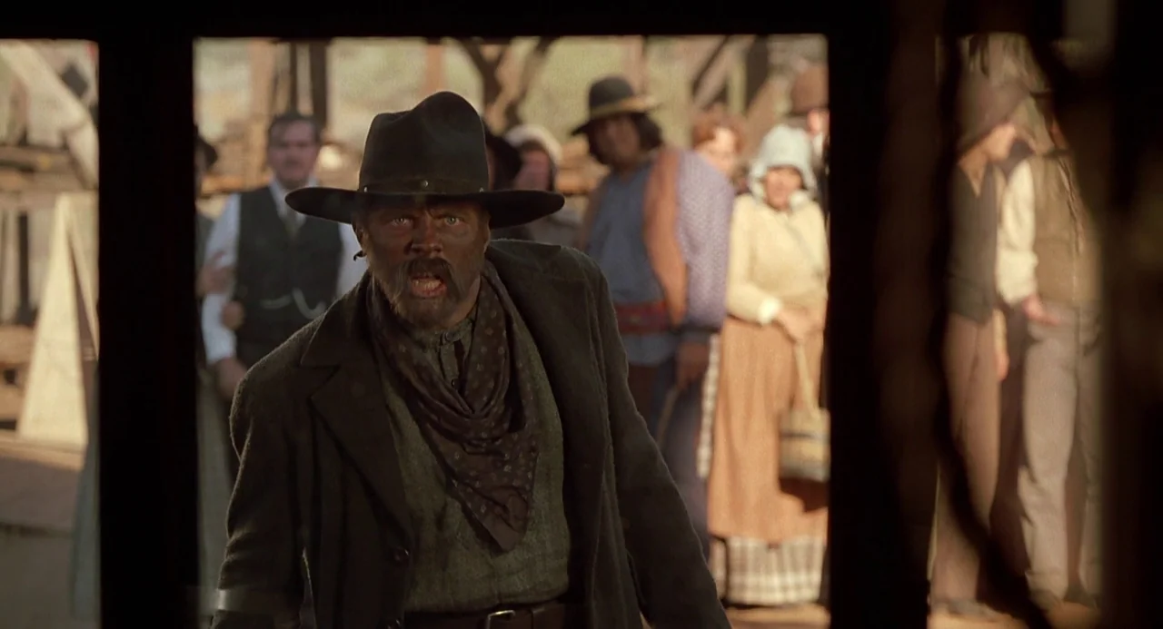Back To The Future Part III (1990) movie still: medium — A dusty outdoor western street scene framed by a dark doorway. A man wearing a cowboy hat…