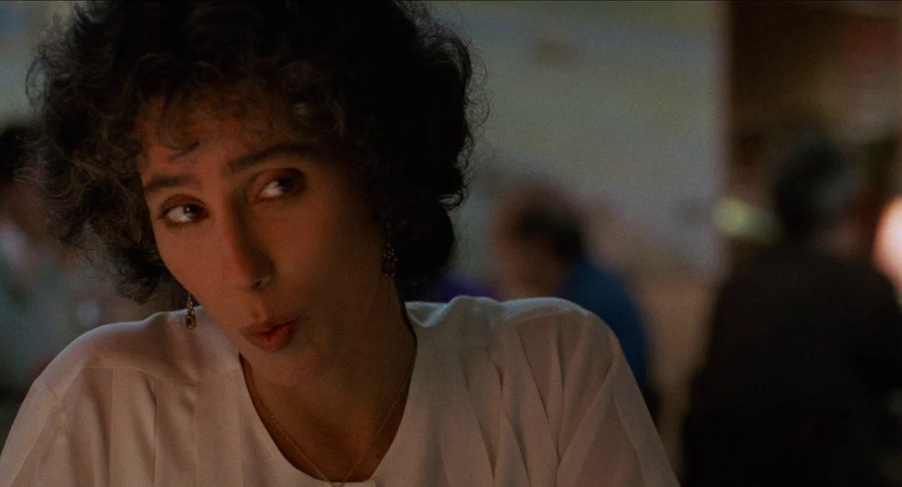 Moonstruck (1987) movie still: closeup — A dimly lit restaurant setting features blurred patrons seated in the dark background. A woman with curly…
