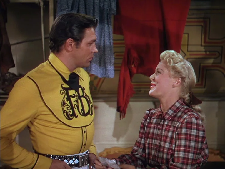 Annie Get Your Gun (1950) movie still: medium — An interior space features clothes hanging on a rack against a pale wall. A man in a yellow shirt…