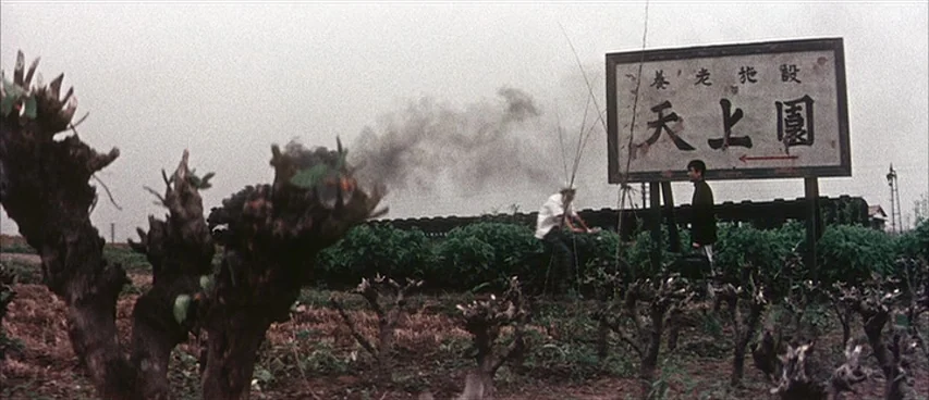 Jigoku (1960) movie still: wide — An open agricultural field with pruned trees features a large weathered signboard in Chinese. One man stands by…