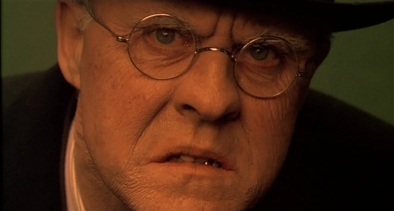 Raising Cain movie still: closeup — A close-up shot focuses on an elderly man's face in a dark room. He stares intensely forward wearing round…