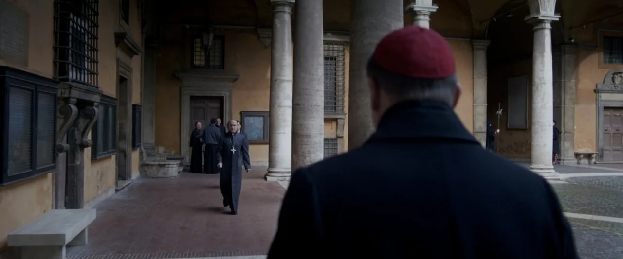 Conclave (2024) movie still: medium — A stone courtyard features tall columns and ochre-colored walls in an institutional setting. A man in black…