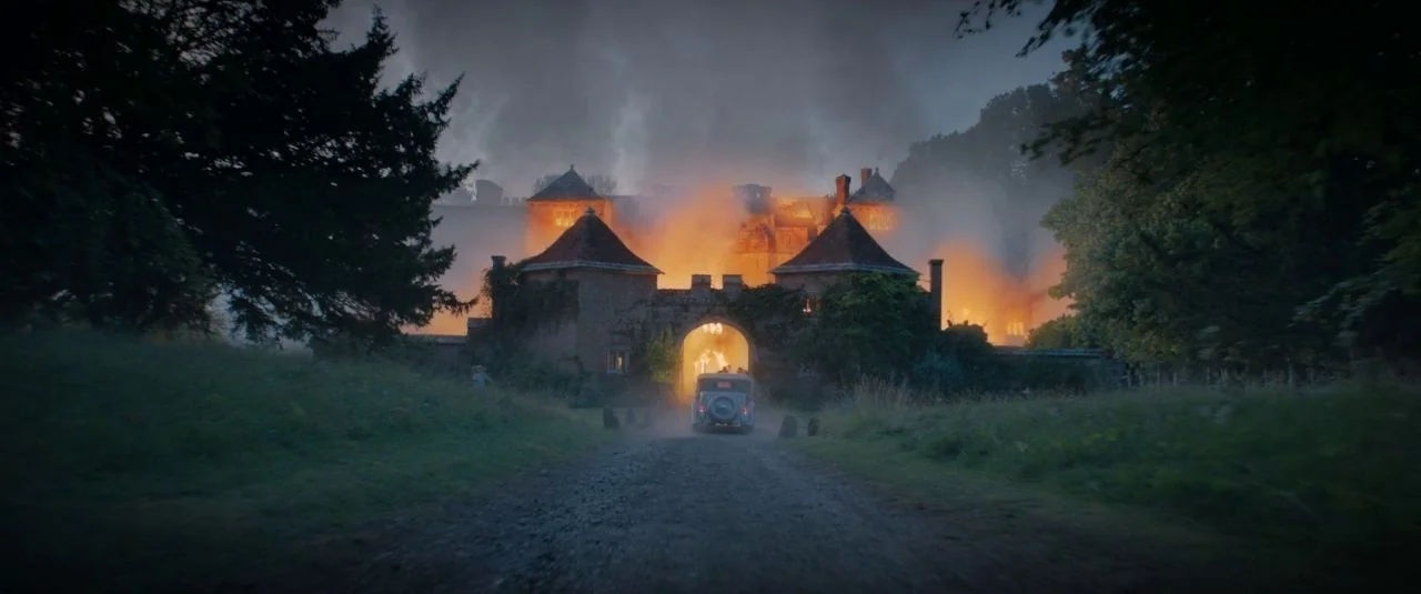 Rebecca movie still: wide — A stone manor house is engulfed in flames with thick plumes of dark smoke. A vintage car drives along a dirt road toward…