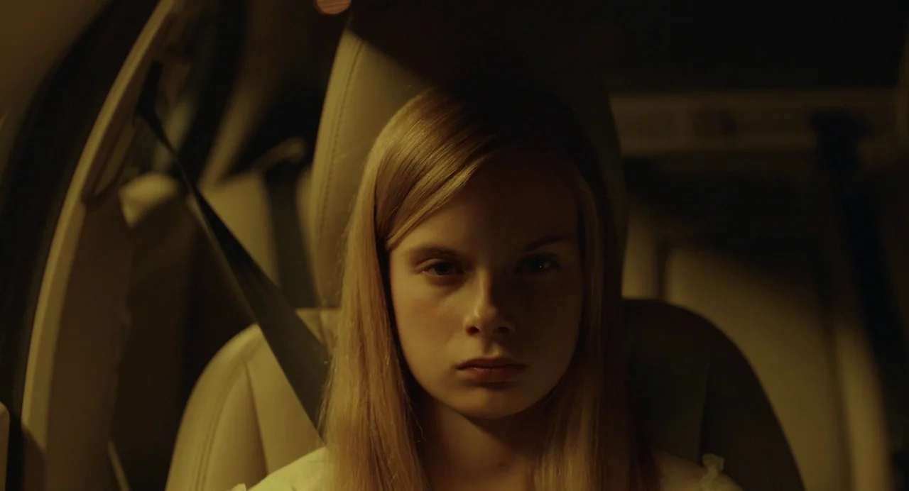 Hatching (2022) movie still: closeup — The interior of a car at night featuring a young woman seated buckled up. She looks straight ahead while…