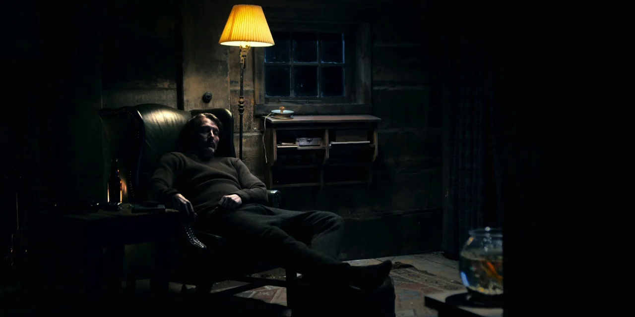 Polar (2019) movie still: wide — A dark wooden room containing a single wingback chair and a small wall shelf. A man in dark clothing sits reclined…