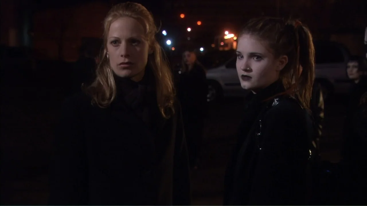O código oculto (2025) movie still: medium — An outdoor parking lot at night with dark surroundings and blurred vehicle lights in back. Two women…