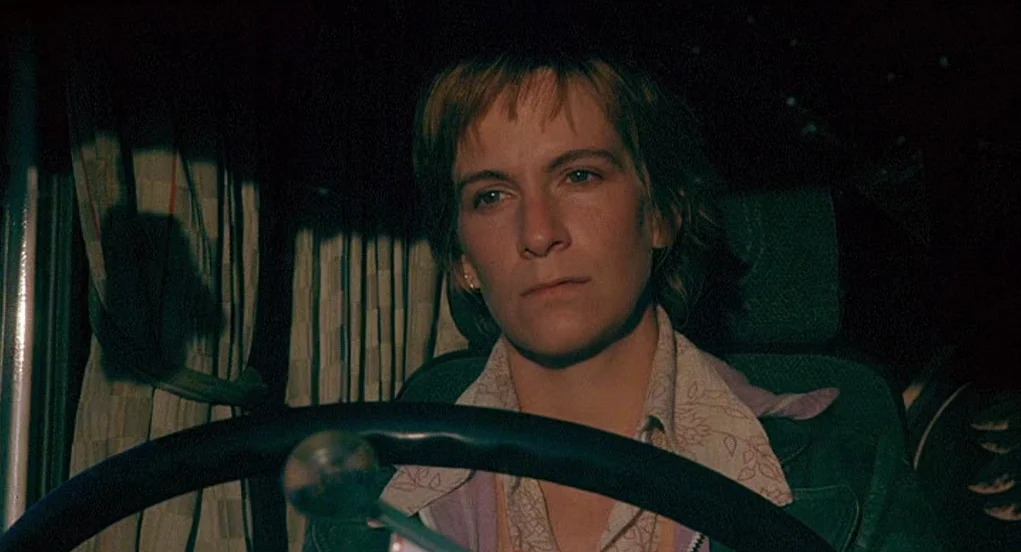 Butterfly Kiss (1995) movie still: medium — A woman sits in the driver's seat of a large truck cab at night. Patterned curtains are visible behind…