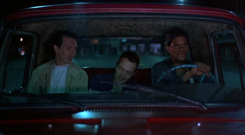 Mystery Train (1989) movie still: medium — Three men sit crowded together inside the front cabin of a vehicle at night. The driver grips the…