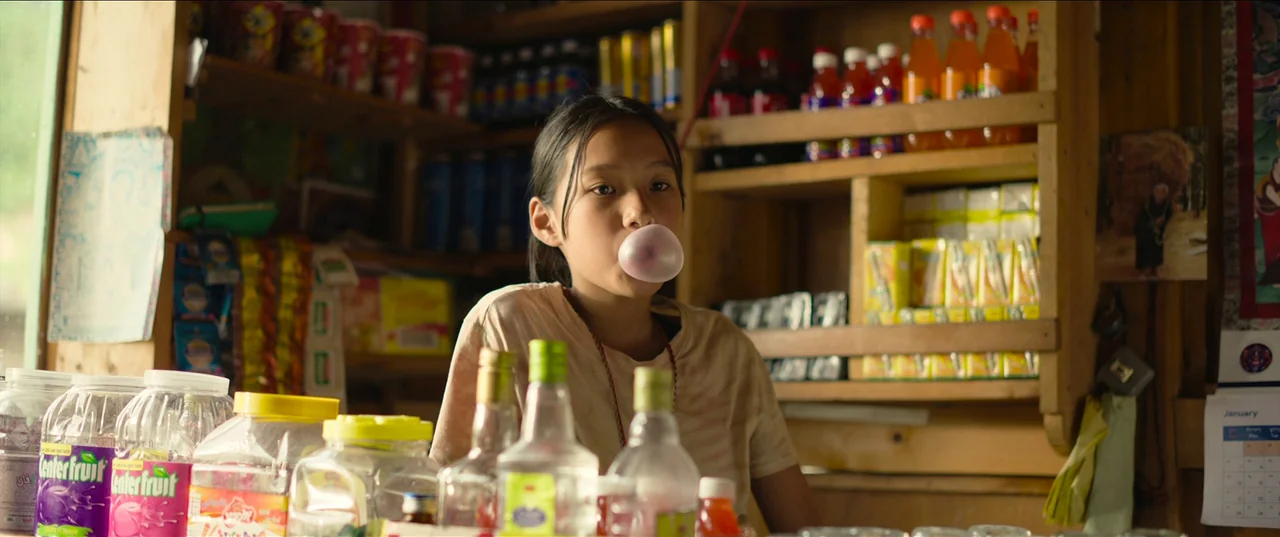 The Monk and The Gun (2023) movie still: medium — A teenage girl stands behind the counter of a cluttered small goods store. She blows a pink bubble…