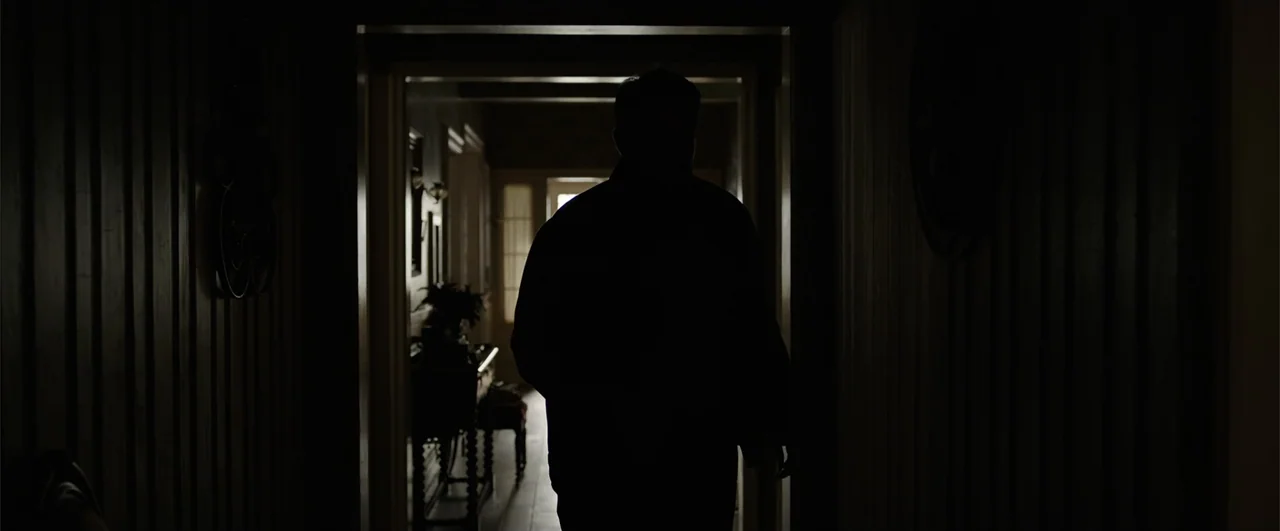 Annabelle: Creation (2017) movie still: medium — A dark interior hallway shows an open doorway leading to a bright room beyond. A lone man is…