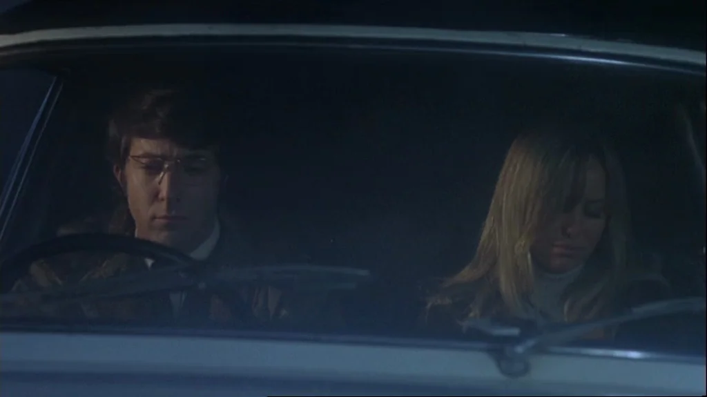 Straw Dogs (1971) movie still: medium — A dark, cramped interior of a vehicle at night with two occupants. The man drives while the woman stares…