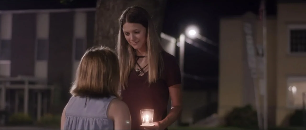 If You're Gone (2019) movie still: medium — A woman stands in an outdoor area at night holding a small candle. She faces a child who has her back to…