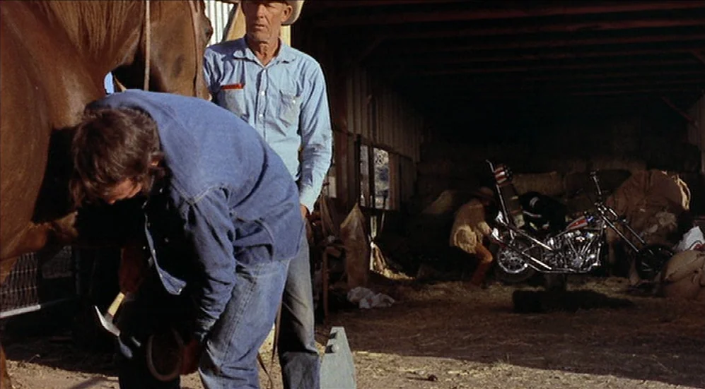 Easy Rider (1969) movie still: medium — A rustic barn interior contains scattered hay and a parked motorcycle in the background. A man works on a…
