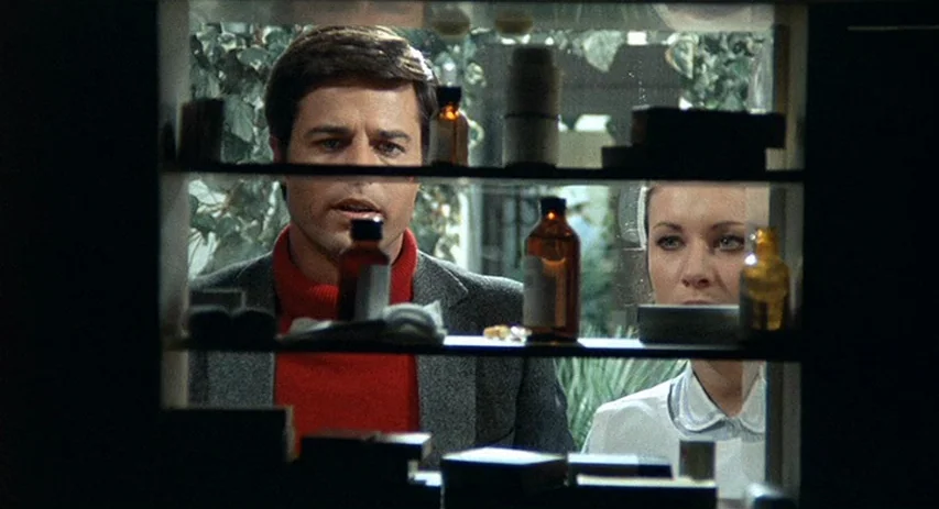 Perversion Story (1969) movie still: medium — A man and woman are visible through the glass shelves of an open cabinet. The pair remains static as…