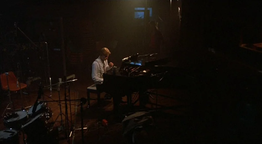 The Coca-Cola Kid (1985) movie still: wide — A dark, cavernous recording studio contains musical equipment and an upright piano. A lone musician…