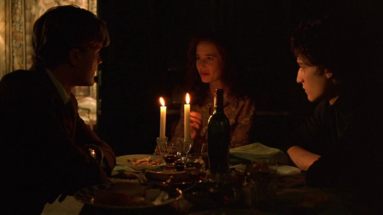 The Dreamers (2003) movie still: medium — A dimly lit room features a table set for dinner with three people. Two men and a woman are seated…