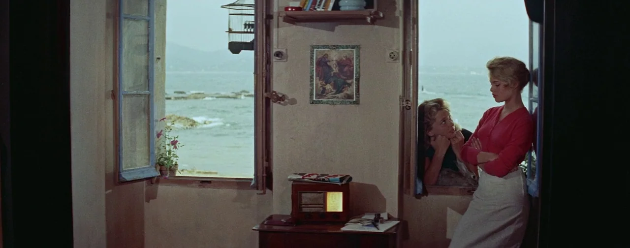 And God Created Woman (1956) movie still: wide — A stone-walled interior room with dual windows overlooking a calm coastal sea. One woman stands in…