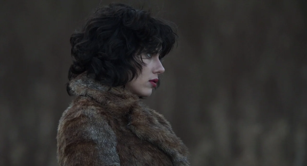 Under The Skin (2013) movie still: closeup — A woman stands in a blurred, dark, and open natural field at dusk. She is looking to the side in a…