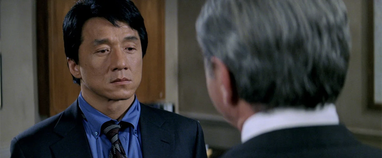 Rush Hour 2 (2001) movie still: medium — An office interior features wood-paneled walls and neutral professional decor. A man in a suit looks toward…
