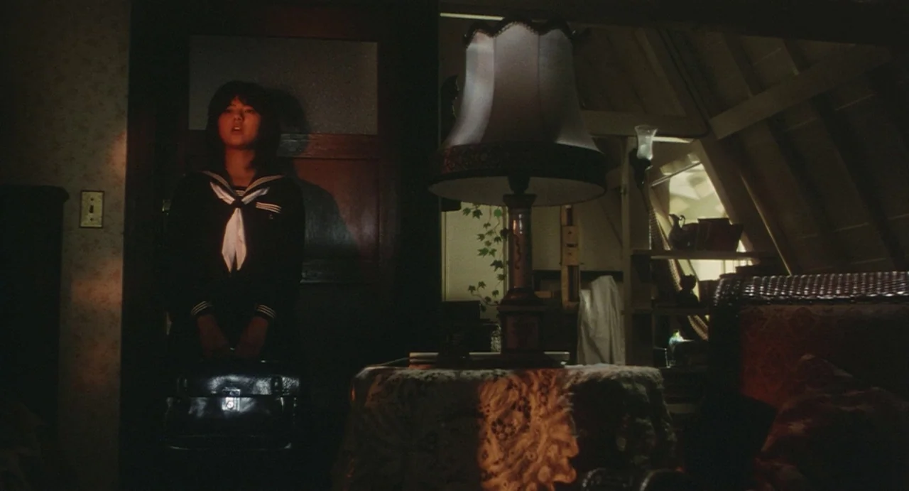 School in the Crosshairs (1981) movie still: medium — An attic space featuring a wooden sloped ceiling and an ornate table lamp. A young girl stands…