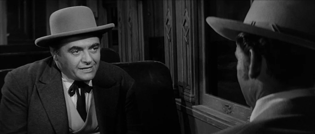 Love Me Tender movie still: medium — A dimly lit train carriage features wood-paneled walls and passenger seating in the background. Two men wearing…