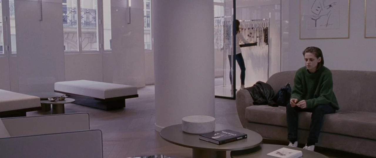 Personal Shopper (2016) movie still: medium — A modern, minimalist clothing boutique with beige furniture and light wood flooring. A woman in a…