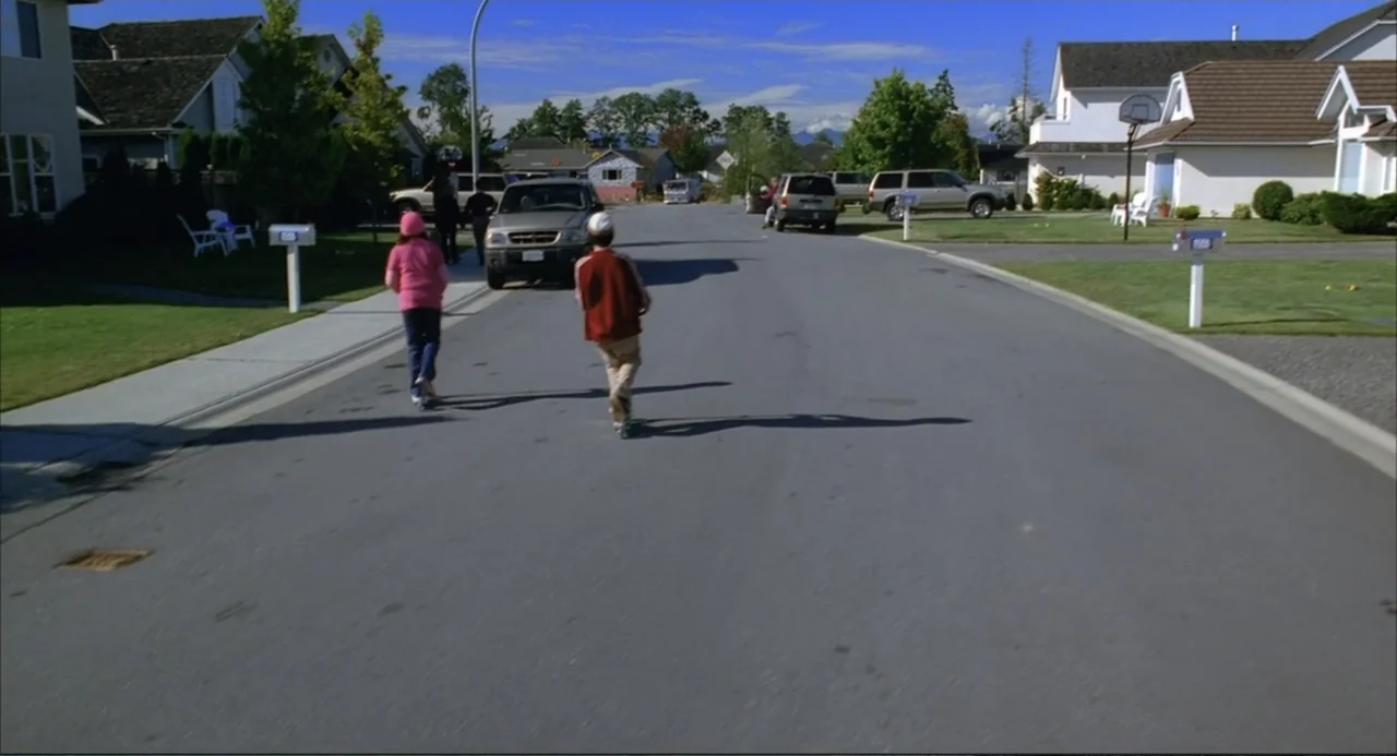Josie and the Pussycats (2001) movie still: wide — A quiet suburban street with houses and parked cars lines both sides of asphalt. Two children in…