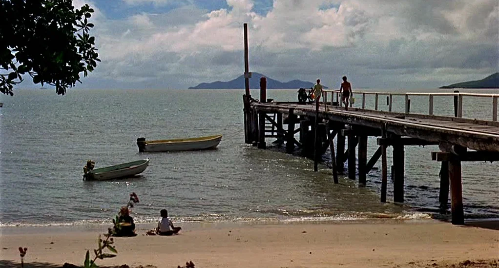 Age of Consent (1969) movie still: wide — A long wooden pier extends into calm seawater under a sky with scattered clouds. Two figures walk along…