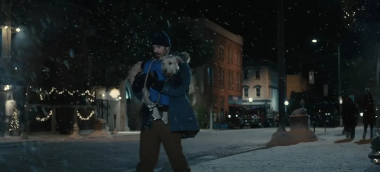 Merv (2025) movie still: fullbody — A snowy street at night lined with buildings and glowing holiday lights. A man walks down the empty road…