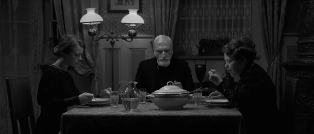 Frantz (2016) movie still: medium — A dimly lit dining room features an old-fashioned double-shaded lamp over a dining table. Two women and one…
