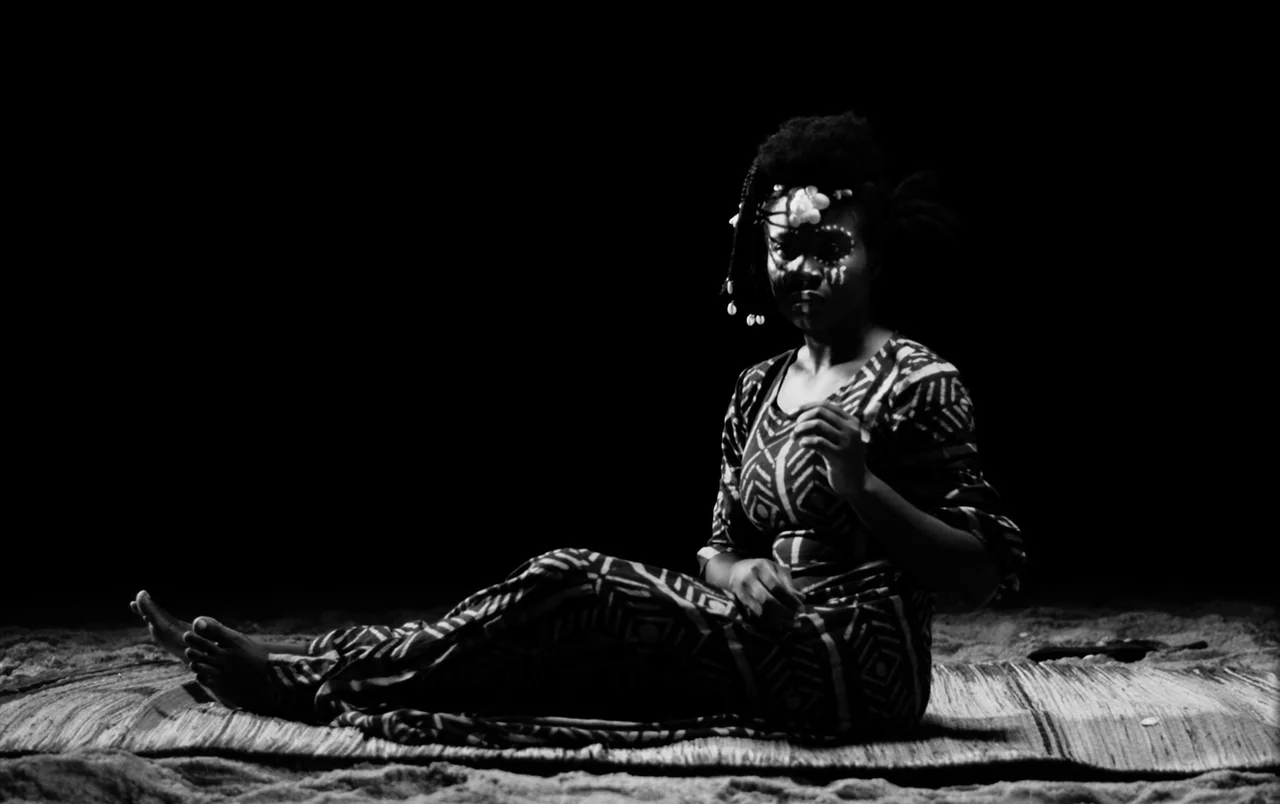 Mami Wata (2023) movie still: fullbody — A dark, empty stage features a woman sitting on a woven floor mat. She wears a patterned dress and…