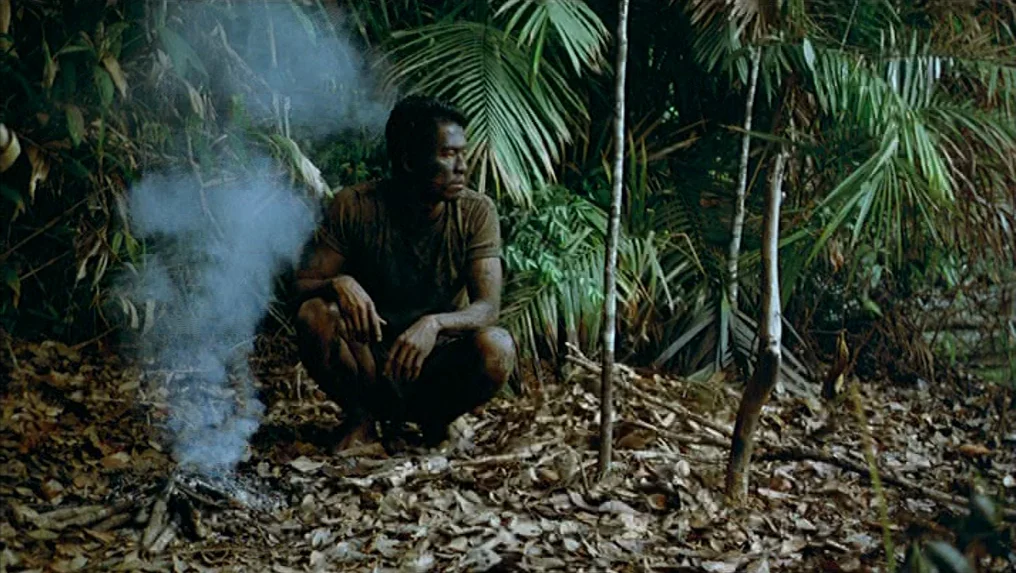 Tropical Malady (2004) movie still: medium — A dense tropical jungle floor covered in dry leaves and lush palm fronds. A man crouches near a small…