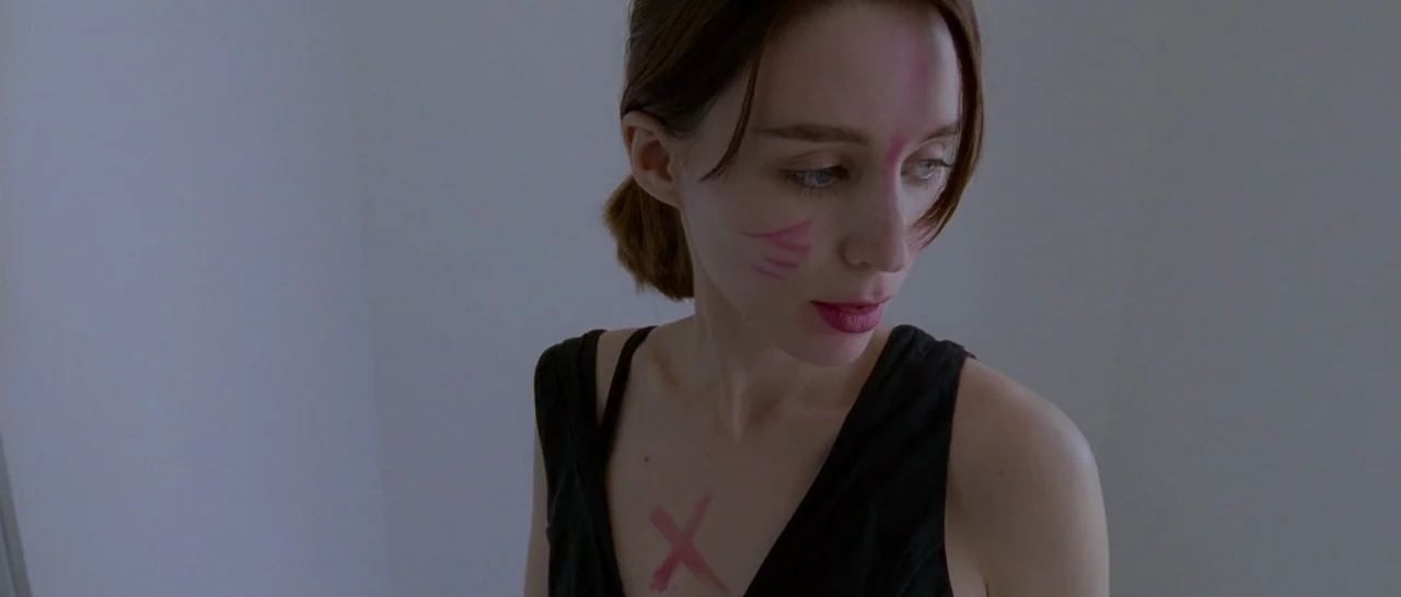 Song to Song (2017) movie still: closeup — A woman stands in front of a plain white wall in a sterile room. She wears a black top and has pink X and…