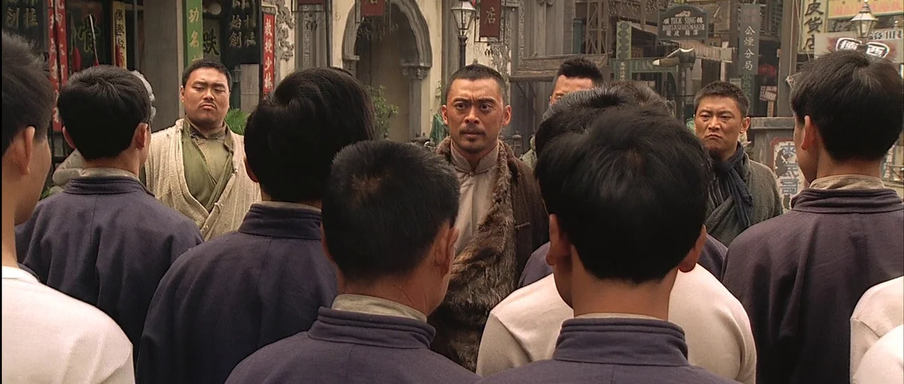Ip Man (2008) movie still: medium — A crowded city street features traditional Chinese shop signs and architectural details. A central man stands in…